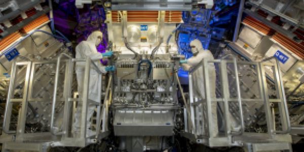 technicians stand in front of large machinery, the target chamber, of National Ignition Facility" title="The target chamber of the National Ignition Facility is contained in the blue sphere at the bottom. Here, technicians performed maintenance of fiber optics.Credit...Jason Laurea/Lawrence Livermore National Laboratory