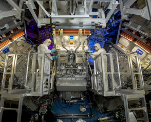 technicians stand in front of large machinery, the target chamber, of National Ignition Facility" title="The target chamber of the National Ignition Facility is contained in the blue sphere at the bottom. Here, technicians performed maintenance of fiber optics.Credit...Jason Laurea/Lawrence Livermore National Laboratory
